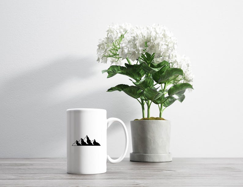 May include: White ceramic mug with a black mountain range design. The mug is sitting on a wooden table next to a potted white flower.