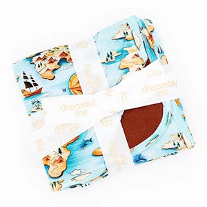 May include: A folded light blue fabric featuring a colorful map print with islands, mountains, palm trees, sailing ships, and sea creatures. A white ribbon with the repeating text dreambly me is wrapped around the fabric, revealing a dark brown interior.