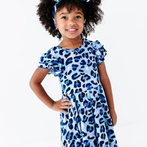May include: A light blue dress featuring a dark blue leopard print pattern with small white star and moon accents. The dress has short ruffled sleeves and a matching headband. The fabric displays a repeating animal print design throughout.