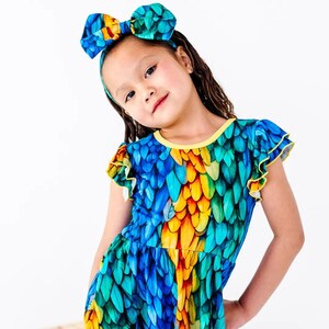 May include: A dress featuring a vibrant feather pattern in shades of blue, teal, and yellow with ruffled sleeves and a yellow neckline trim. The dress is paired with a matching headband that has a large bow, both displaying the same colorful feather print.