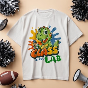 Peut inclure: T-shirt blanc avec un dinosaure de dessin animé tenant un marqueur, avec le texte "CLASS IS MY LAB" en orange, jaune et vert. Le dinosaure est vert avec des accents orange, sur des éclaboussures de peinture colorées.
