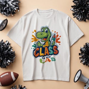 Peut inclure: T-shirt blanc avec un dinosaure de dessin animé tenant un crayon, avec le texte "CLASS IS MY LAB". Le dinosaure est vert et orange, avec des yeux bleus. Le t-shirt a un col rond.