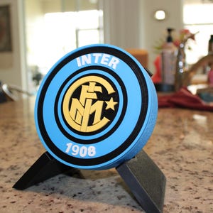 May include: A blue and black Inter Milan logo displayed on a stand. The logo features the word "INTER" in white, the year "1908", and a yellow and black emblem. The stand is black.