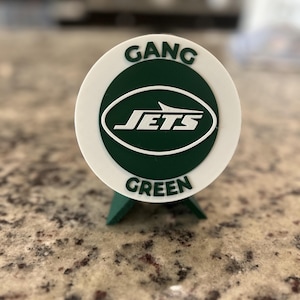May include: A circular white and dark green plastic sign featuring the text GANG GREEN in dark green lettering and the white Jets logo in the center. The sign is supported by a small dark green stand attached to the bottom of the circular frame.