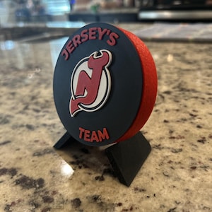 May include: A circular black and red hockey puck-style display piece featuring the New Jersey Devils logo in the center. The text "JERSEY'S" is arched above the logo and "TEAM" is below it in red lettering. It rests on a black stand.