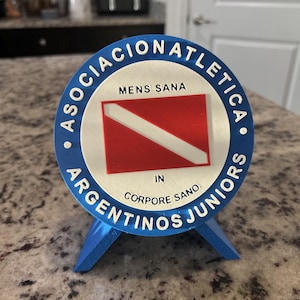 May include: A circular 3D printed emblem featuring the text ASOCIACION ATLETICA ARGENTINOS JUNIORS in blue, with MENS SANA IN CORPORE SANO in black, and a central red and white flag design. The emblem is supported by a blue stand.