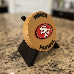 May include: A circular gold-colored plaque featuring the San Francisco 49ers logo in red, white, and black, with the black text NINER FAITHFUL printed above and below the logo, supported by a black stand.