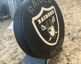 Puede incluir: Una placa circular negra con el logotipo de los Raiders en blanco y negro, con el texto RAIDER NATION grabado en gris. La placa está montada sobre un pequeño soporte negro inclinado y presenta un acabado de superficie texturizado.