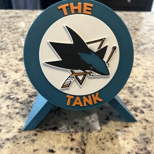 May include: A circular teal display stand featuring a white center disc with a black and teal shark logo biting a hockey stick. The text THE TANK is embossed in orange letters at the top and bottom of the teal border. The stand has two angled supports.