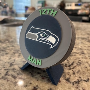 May include: A circular 3D printed display piece featuring a dark blue center with a white and grey bird logo. The grey border has the text 12TH in green at the top and MAN in green at the bottom. The object rests on a dark blue angled stand.
