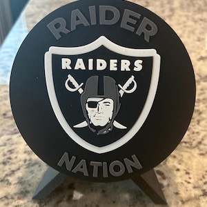 May include: A circular black plaque featuring the Raiders logo in the center, with the word RAIDER arched above and NATION arched below in grey lettering. The shield logo displays a football player wearing an eye patch and two crossed swords.