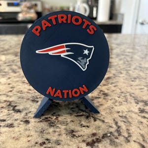 May include: A circular dark blue display plaque featuring the red text "PATRIOTS" arched at the top and "NATION" at the bottom. In the center is the New England Patriots logo, a stylized profile of a person wearing a tricorne hat in red, white, and blue.