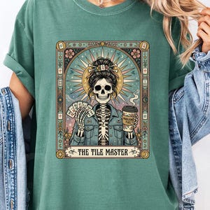 Pode incluir: Camiseta verde com um design de carta de tarô. O design apresenta um esqueleto segurando cartas e uma xícara de café, com o texto "THE TILE MASTER" em uma moldura decorativa. A camiseta tem mangas enroladas.