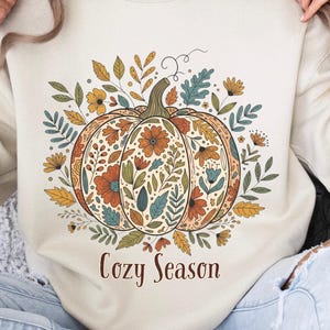 Può includere: Felpa color crema con un design dettagliato di zucca con accenti floreali e di foglie arancioni, gialle e verde acqua. Le parole "Cozy Season" sono stampate in marrone sotto la zucca. La felpa ha una vestibilità ampia.