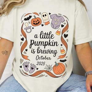 Può includere: T-shirt color crema con grafica a tema Halloween. Il design include una zucca, un fantasma, vestiti per bambini e la scritta "a little pumpkin is brewing October 2025". Il design è incorniciato da un nastro arancione.