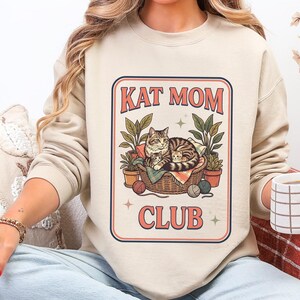 May include: Beige sweatshirt with a graphic that reads "Kat Mom Club." The graphic features a cat in a basket with plants and yarn. The sweatshirt is worn by a person sitting on a couch.
