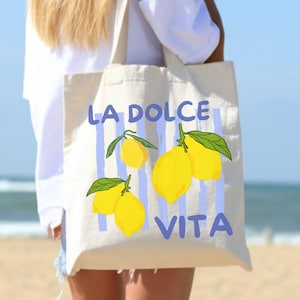 A woman is standing on a beach, carrying a tote bag with a design featuring lemons and the text "LA Dolce Vita".