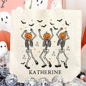 Puede incluir: Bolso tote de lona color crema con un diseño de Halloween. Presenta tres esqueletos bailando con cabezas de calabaza, murciélagos negros y estrellas. El nombre "KATHERINE" está impreso en la bolsa. El fondo incluye una calabaza, fantasmas y bolas de discoteca.