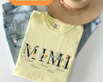 May include: A yellow t-shirt with the word "MIMI" in black letters surrounded by floral designs. The names "Noah, Thomas, Emma, Olivia, James" are written in black below the letters.