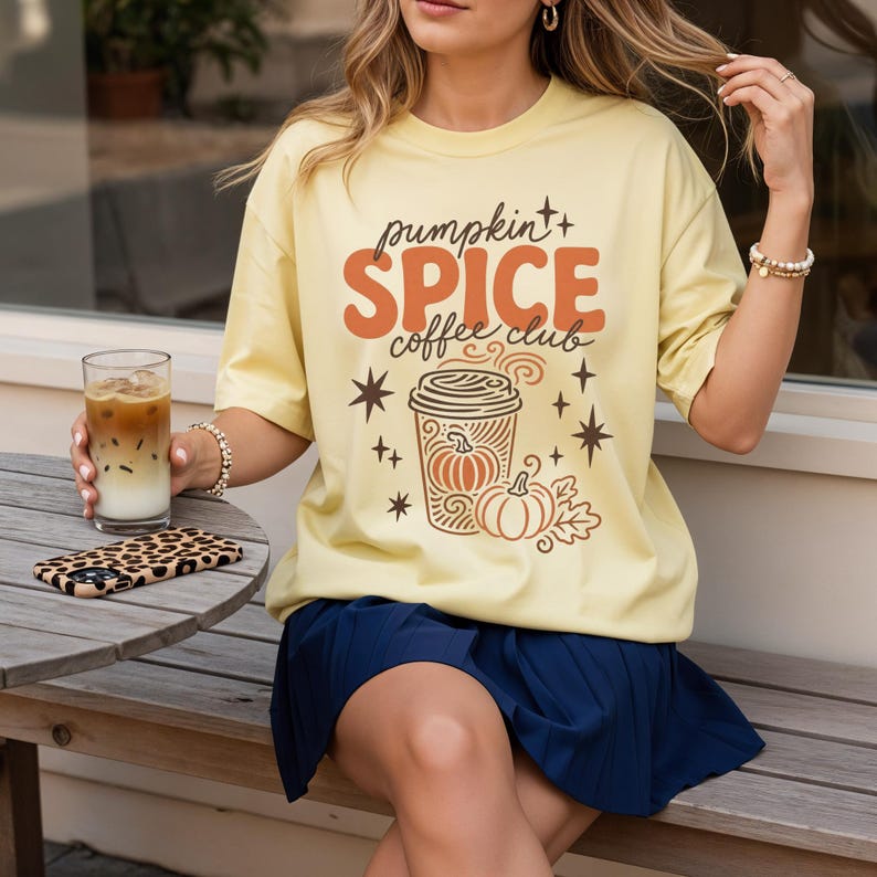 A woman wearing a yellow t-shirt with a pumpkin spice coffee club design is sitting at a table with a glass of iced coffee and a leopard print phone case.