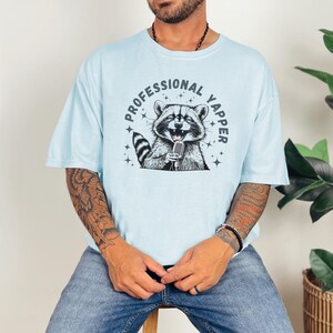a man sitting on a stool wearing a shirt with a raccoon on it