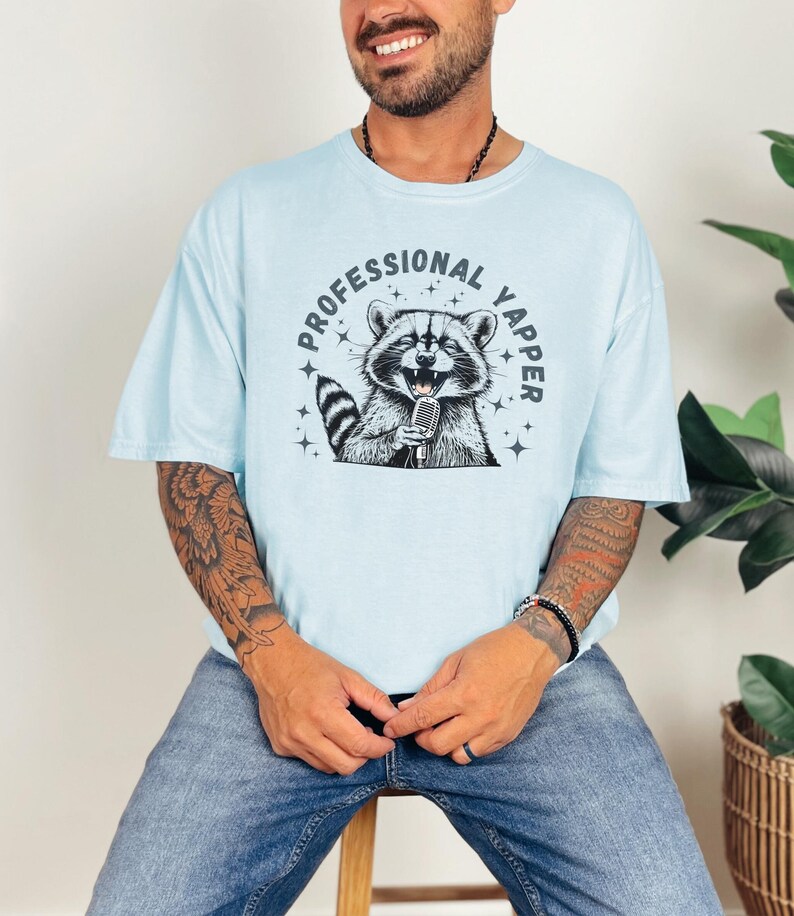 a man sitting on a stool wearing a shirt with a raccoon on it