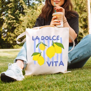 A woman sitting on the grass, holding a drink and a bag with a graphic design featuring lemons.