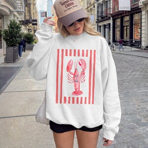 White crewneck sweatshirt with a watercolor lobster graphic on red and white stripes.