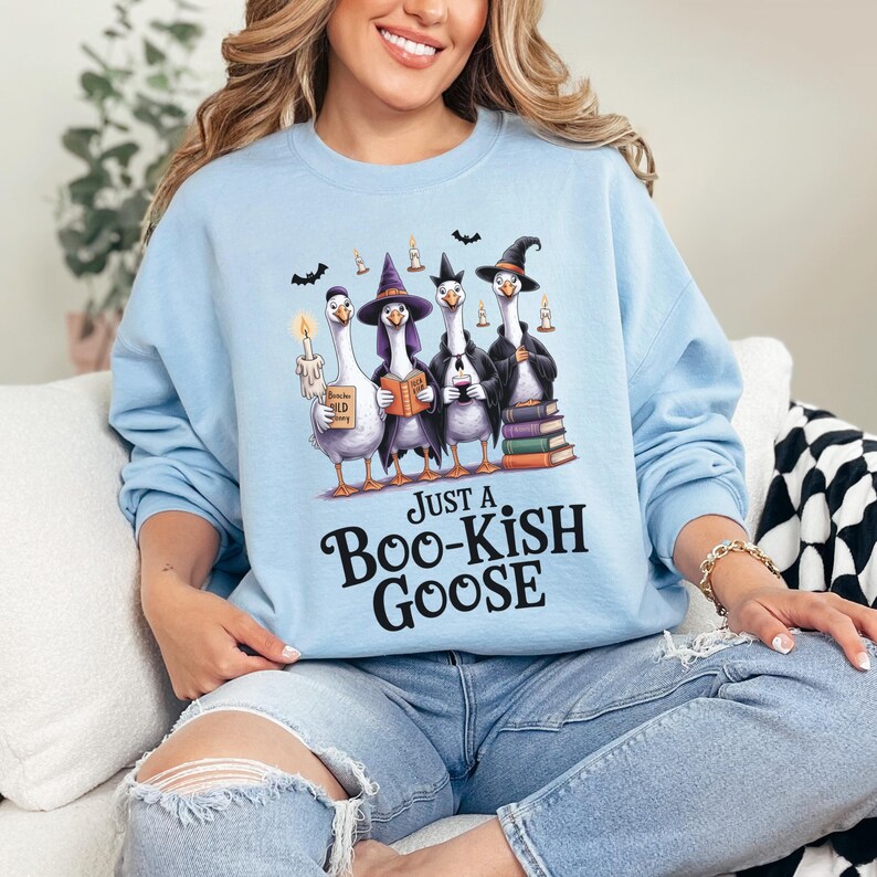 A woman wearing a light blue sweatshirt with a graphic of four cartoon geese dressed as witches and a book titled "Just a Boo-Kish Goose".