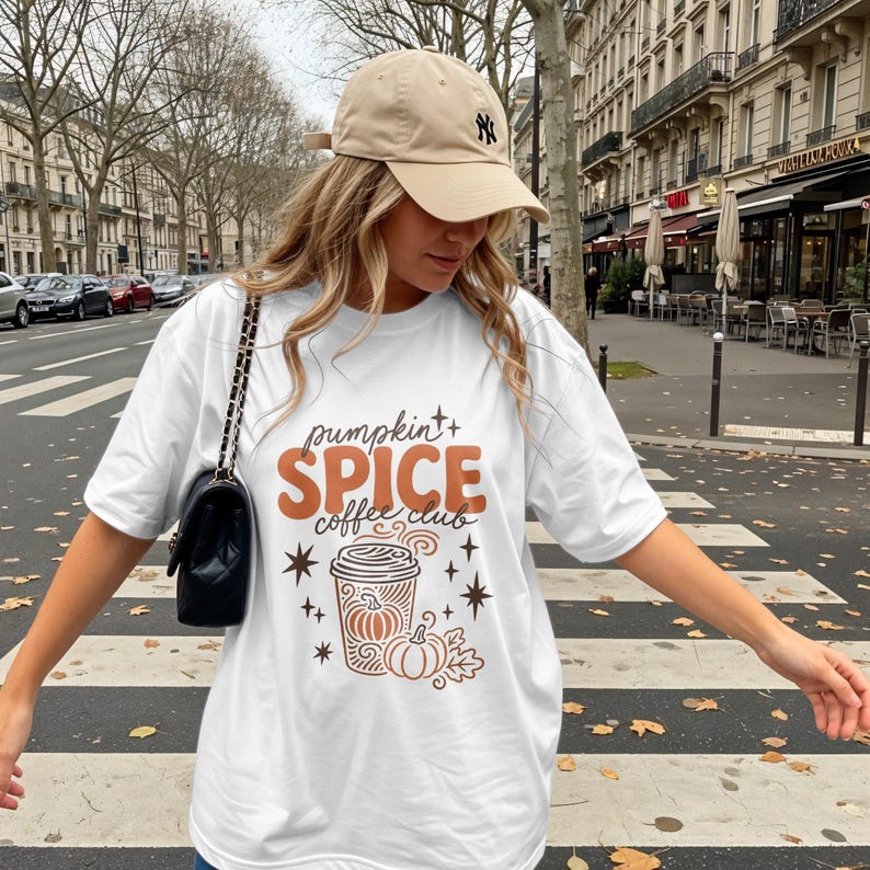 White t-shirt with "Pumpkin Spice Coffee Club" text and autumnal graphics on a street.