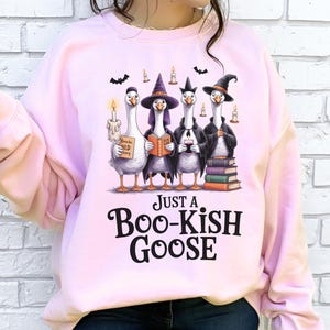 A woman wearing a pink sweatshirt with a graphic design of four cartoon geese dressed in witch hats and holding various objects, including a candle, a book, and a sign that says "Just a Boo-Kish Goose".