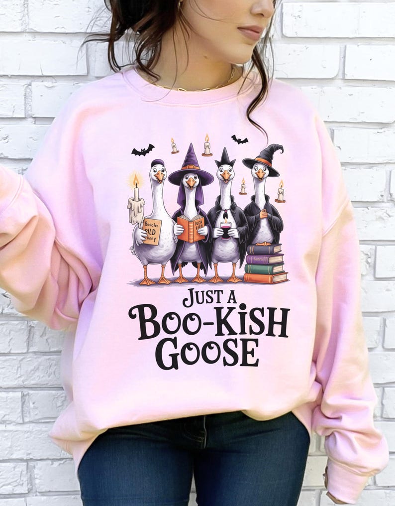 A woman wearing a pink sweatshirt with a graphic design of four cartoon geese dressed in witch hats and holding various objects, including a candle, a book, and a sign that says "Just a Boo-Kish Goose".