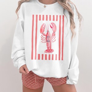 White crewneck sweatshirt with watercolor lobster graphic and red stripes.