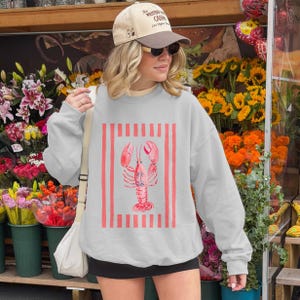 Woman wearing a light gray sweatshirt with a pink lobster graphic and red stripes.