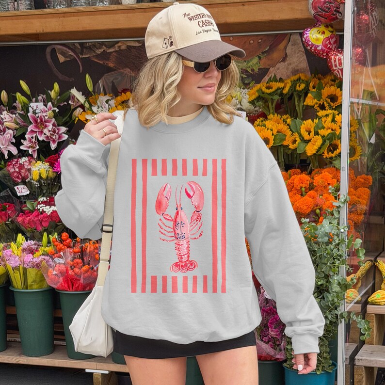 Woman wearing a light gray sweatshirt with a pink lobster graphic and red stripes.