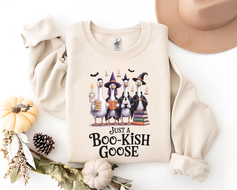A beige sweater with a Halloween-themed graphic of four geese dressed as witches and a boomerang, along with the text "Just a Boo-Kish Goose" printed on it.