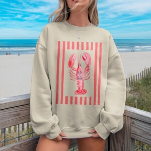 Cream crewneck sweatshirt with a pink lobster design on a beach background.