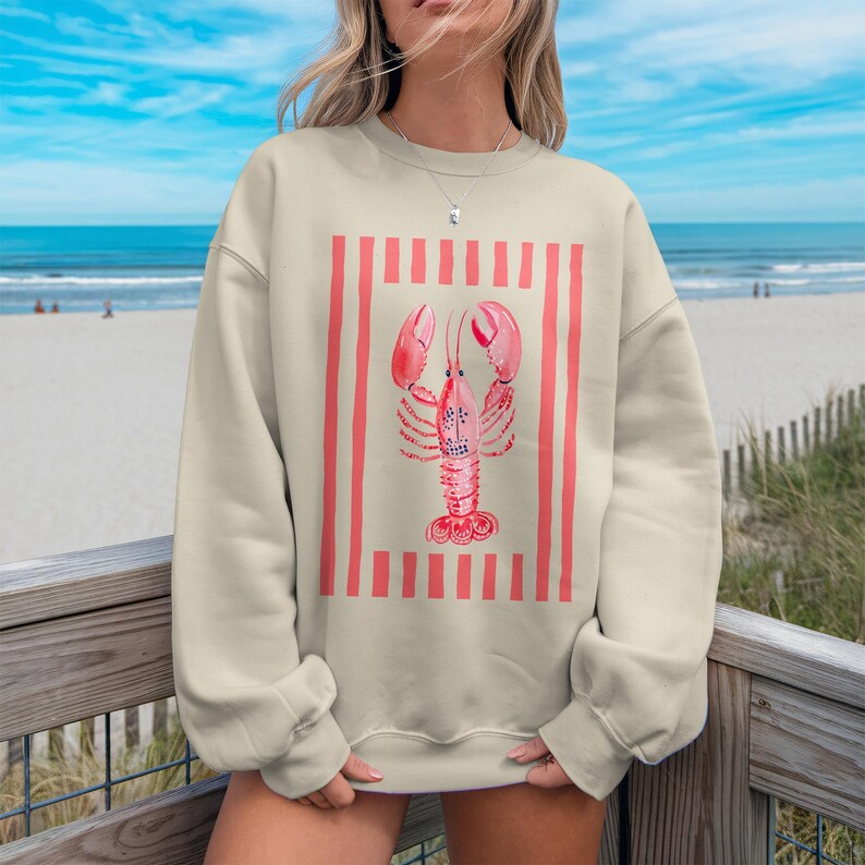 Cream crewneck sweatshirt with a pink lobster design on a beach background.