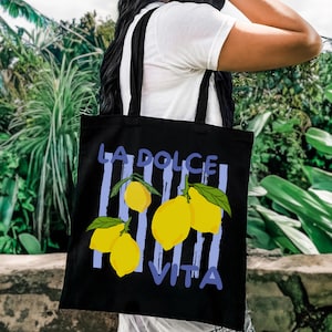 A person is carrying a black tote bag with a graphic design featuring yellow lemons and the text "LA DOLCE VITA" printed on it.