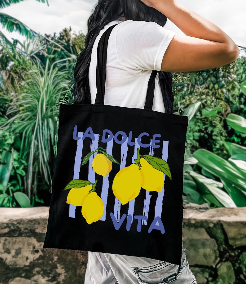 A person is carrying a black tote bag with a graphic design featuring yellow lemons and the text "LA DOLCE VITA" printed on it.