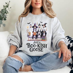 A woman wearing a gray sweatshirt with a graphic of four cartoon geese dressed in witch costumes and holding candles.