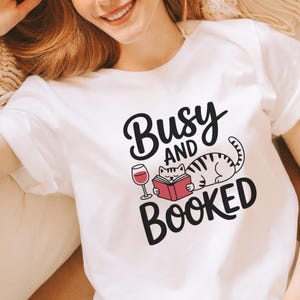 Puede incluir: Camiseta blanca con el texto "Busy and Booked" en una fuente caprichosa. El diseño incluye un gato de dibujos animados leyendo un libro, una copa de vino y un libro rojo. La camiseta es de estilo casual y cómodo.