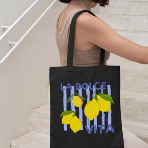 A woman is walking down a staircase while carrying a black tote bag with a graphic design featuring lemons and the text "LA DOLCE VITA".