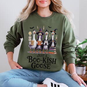 A woman wearing a green sweatshirt with a graphic of four cartoon geese dressed in witch's hats and holding books, along with the text "Just a Boo-Kish Goose".