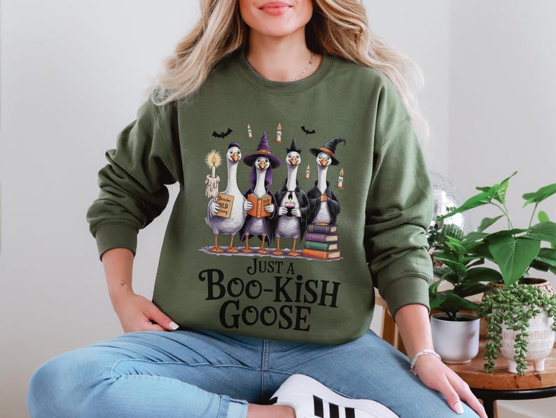 A woman wearing a green sweatshirt with a graphic of four cartoon geese dressed in witch's hats and holding books, along with the text "Just a Boo-Kish Goose".