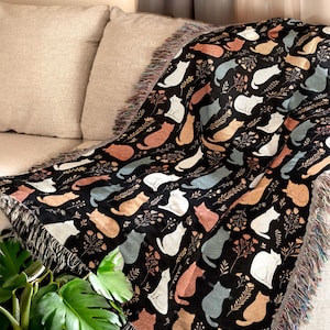 Könnte beinhalten: Schwarze Decke mit einem sich wiederholenden Muster aus stilisierten Katzen in Weiß, Orange und Grau. Die Decke hat einen Fransenrand und ist über einem beigen Sofa drapiert. Größe: ca. 150 cm x 130 cm.