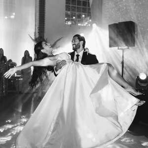 Puede incluir: Imagen en blanco y negro de una celebración de boda. Una persona con un vestido blanco es sostenida por otra con un traje negro, ambos sonriendo. Los focos iluminan la escena, creando un ambiente festivo.