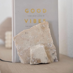 Może przedstawiać: Jasnoszara książka ze złotymi literami z napisem "GOOD VIBES" i "HOW SELF-LOVE IS THE KEY". Przed książką znajdują się dwa kawałki jasnego, szorstkiego kamienia.