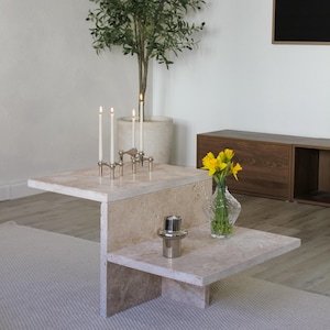 Könnte beinhalten: Ein weißer Travertin-Couchtisch mit einem einzigartigen asymmetrischen Design. Der Tisch hat zwei Ebenen und verfügt über einen silbernen Kerzenhalter mit drei brennenden Kerzen und eine silberne Vase mit gelben Blumen.