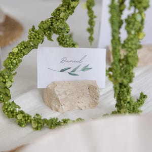 Könnte beinhalten: Eine Platzkarte mit dem Namen "Daniel" in eleganter Schrift, verziert mit einem Zweig Blättern, liegt auf einem kleinen, strukturierten Stein. Die Karte ist von grünen floralen Elementen umgeben, was eine natürliche, elegante Ästhetik schafft. Ideal für Hochzeiten oder Veranstaltungen.